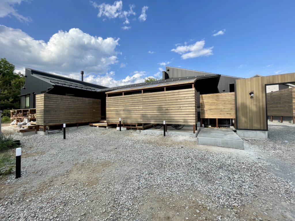 [山梨県八ヶ岳南麓北杜市長坂に建築したパッシブデザインの自然素材の家　建築設計施工事例] 　自然素材の家　別荘　注文住宅専門の建築設計事務所と建てるソーラーシステムそよ風で床暖房するサステナブルな環境共生住宅 平屋のコの字型間取りプラン 画像は駐車スペースの木の羽目板仕上げのゲートと木の塀で目隠しした駐輪場 奥側にガルバリウム仕上げの家があります