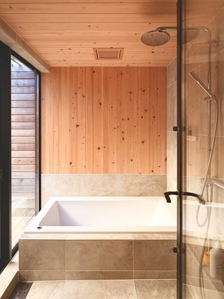 神奈川県鎌倉市西鎌倉に建築したL字型間取り設計の自然素材の家。夏や梅雨時は湿気や熱を排気し、冬は太陽熱で温めた外気を床下に送り換気をしながら床暖房するソーラーシステムそよ風で家中足元から暖かくします。壁天井は消臭調湿効果が高い珪藻土、床は桧フローリングなど天然素材仕上げ。自然素材とソーラーシステムそよ風の相乗効果で湿気が少なく夏涼しく冬暖かいパッシブデザインの環境共生住宅です。 全室収納は造り付けで作成したのでスッキリものが片付き使い勝手と住み心地がいいサステナブルハウス。 画像は桧の羽目板と６００角タイルで仕上げた1.２５坪サイズの浴室。床暖房で冬も暖かく、夜は窓を開け木の塀で目隠しした坪庭の植栽をライトアップして桧の香りとぬくもりに癒され露天風呂気分で入浴できます。