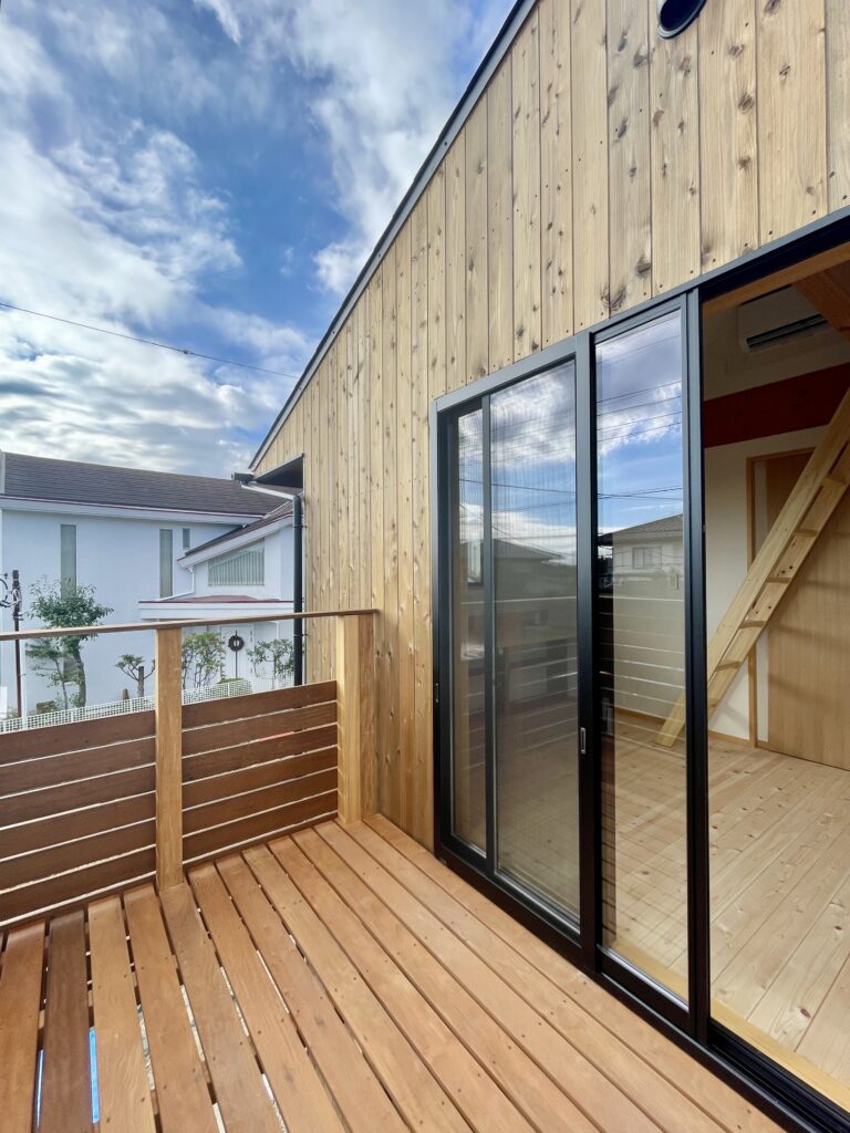 神奈川県鎌倉市西鎌倉に建築したL字型間取り設計の自然素材の家。夏や梅雨時は湿気や熱を排気し、冬は太陽熱で温めた外気を床下に送り換気をしながら床暖房するソーラーシステムそよ風で家中足元から暖かくします。壁天井は消臭調湿効果が高い珪藻土、床は桧フローリングなど天然素材仕上げ。自然素材とソーラーシステムそよ風の相乗効果で湿気が少なく夏涼しく冬暖かいパッシブデザインの環境共生住宅です。 全室収納は造り付けで作成したのでスッキリものが片付き使い勝手と住み心地がいいサステナブルハウス。 画像は多目的室に隣接したウッドデッキのバルコニー。道路側からは木のスリット塀で目隠しされプライバシーを確保しながら湘南海岸と鎌倉山が見えます。