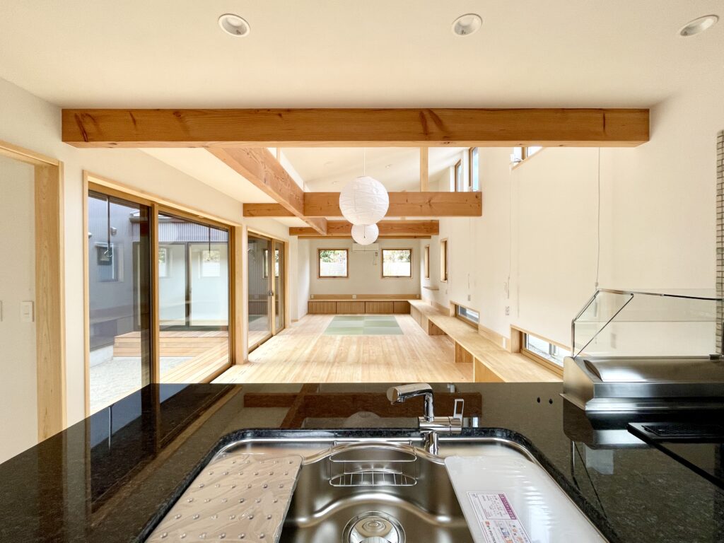 静岡県東部沼津市に建築したコの字型間取り設計の平家の自然素材の家。夏や梅雨時は湿気や熱を排気し、冬は太陽熱で温めた外気を床下に送り換気をしながら床暖房するソーラーシステムそよ風で家中足元から暖かくします。壁天井は消臭調湿効果が高い珪藻土、床は桧フローリングなど天然素材仕上げ。自然素材とソーラーシステムそよ風の相乗効果で湿気が少なく夏涼しく冬暖かいパッシブデザインの環境共生住宅です。 全室収納は造り付けで作成したのでスッキリものが片付き使い勝手と住み心地がいいサステナブルハウス。 画像はキッチン側から見た拭き受けのリビング。東西南北と高窓からの通風と採光で明るく風通しのいいリビングです。