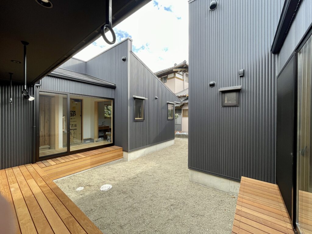 静岡県東部沼津市に建築したコの字型間取り設計の平家の自然素材の家。夏や梅雨時は湿気や熱を排気し、冬は太陽熱で温めた外気を床下に送り換気をしながら床暖房するソーラーシステムそよ風で家中足元から暖かくします。壁天井は消臭調湿効果が高い珪藻土、床は桧フローリングなど天然素材仕上げ。自然素材とソーラーシステムそよ風の相乗効果で湿気が少なく夏涼しく冬暖かいパッシブデザインの環境共生住宅です。 全室収納は造り付けで作成したのでスッキリものが片付き使い勝手と住み心地がいいサステナブルハウス。 画像はコの字型設計プランの中庭。ウッドデッキの縁側。外壁はガルバリウム仕上げ。