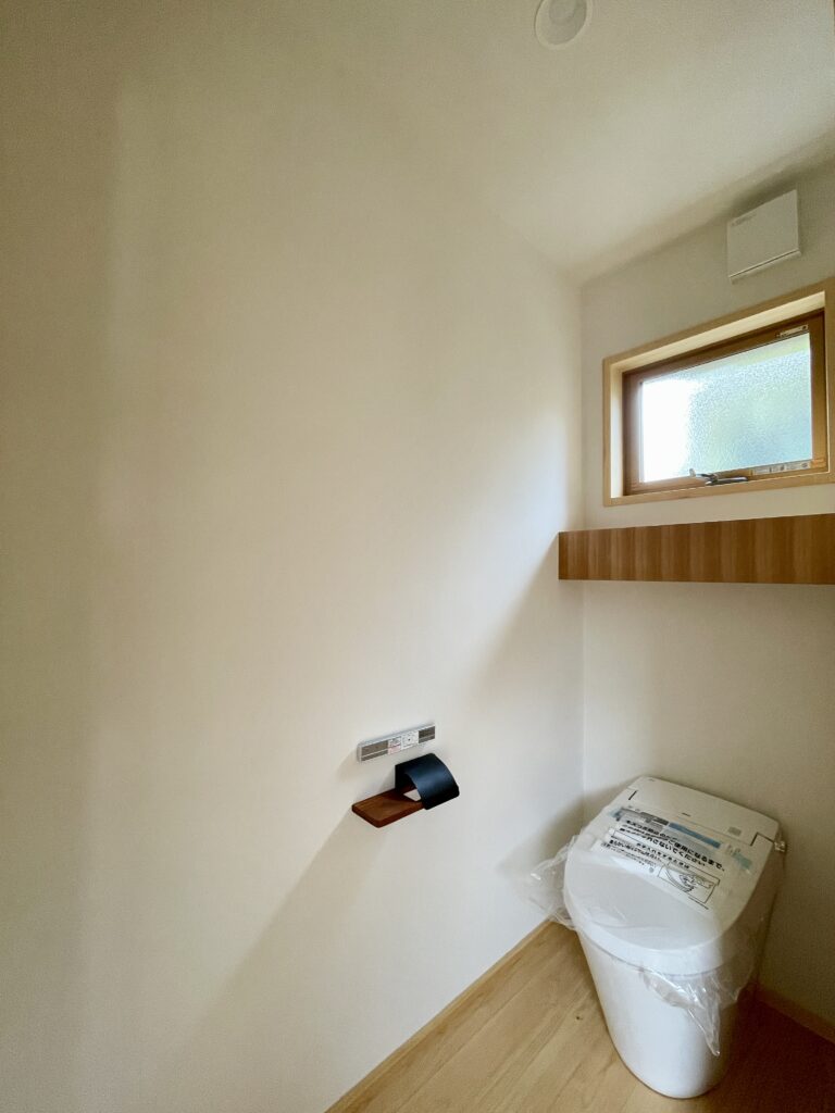 静岡県東部沼津市に建築したコの字型間取り設計の平家の自然素材の家。夏や梅雨時は湿気や熱を排気し、冬は太陽熱で温めた外気を床下に送り換気をしながら床暖房するソーラーシステムそよ風で家中足元から暖かくします。壁天井は消臭調湿効果が高い珪藻土、床は桧フローリングなど天然素材仕上げ。自然素材とソーラーシステムそよ風の相乗効果で湿気が少なく夏涼しく冬暖かいパッシブデザインの環境共生住宅です。 全室収納は造り付けで作成したのでスッキリものが片付き使い勝手と住み心地がいいサステナブルハウス。 画像はトイレットペーパー収納棚があるトイレ。