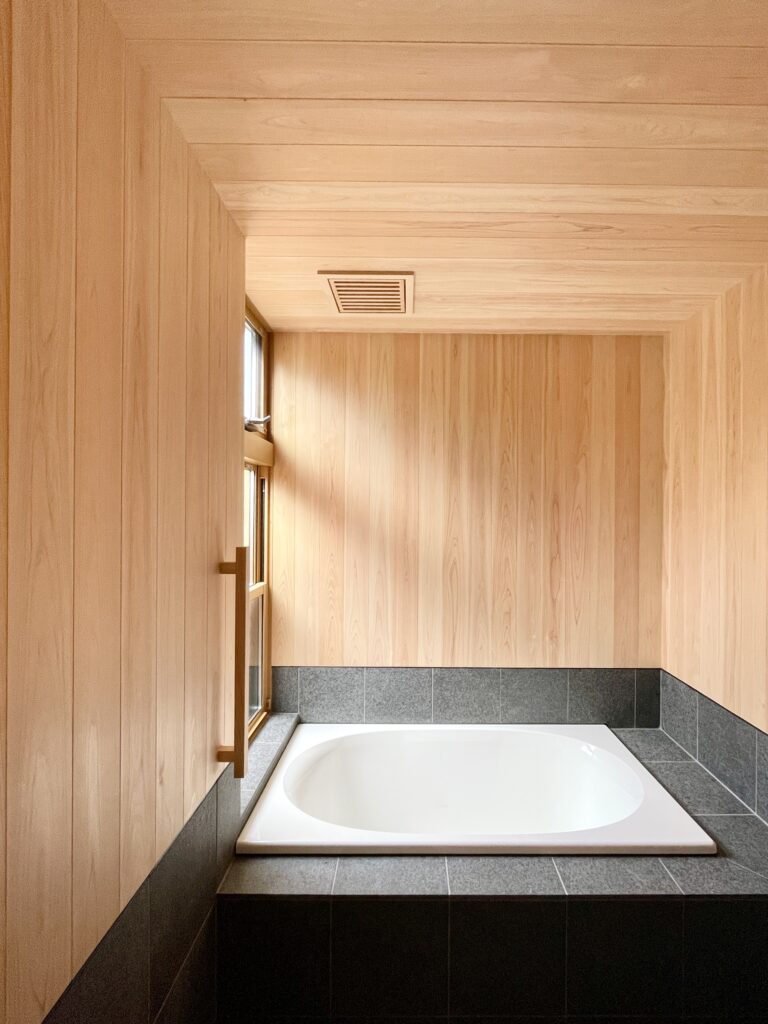 静岡県東部沼津市に建築したコの字型間取り設計の平家の自然素材の家。夏や梅雨時は湿気や熱を排気し、冬は太陽熱で温めた外気を床下に送り換気をしながら床暖房するソーラーシステムそよ風で家中足元から暖かくします。壁天井は消臭調湿効果が高い珪藻土、床は桧フローリングなど天然素材仕上げ。自然素材とソーラーシステムそよ風の相乗効果で湿気が少なく夏涼しく冬暖かいパッシブデザインの環境共生住宅です。 全室収納は造り付けで作成したのでスッキリものが片付き使い勝手と住み心地がいいサステナブルハウス。 画像は桧の羽目板と３００角御影石で仕上げた1.２５坪サイズの浴室。床暖房で冬も暖かく、夜は窓を開け木の塀で目隠しした坪庭の植栽をライトアップして桧の香りとぬくもりに癒され露天風呂気分で入浴できます。