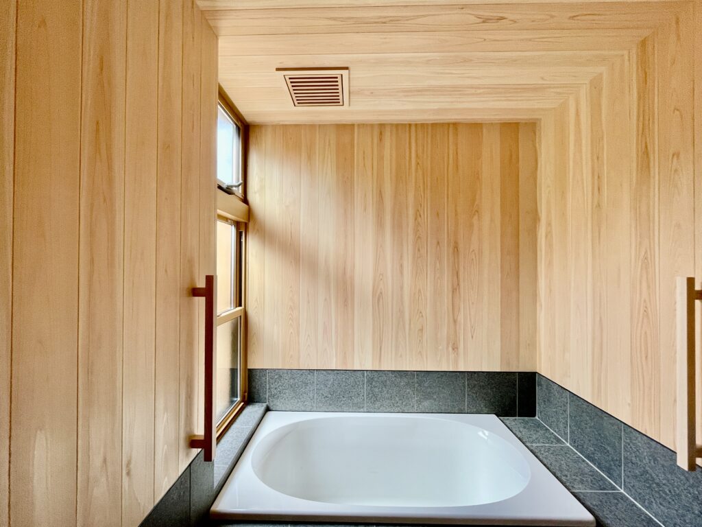 静岡県東部沼津市に建築したコの字型間取り設計の平家の自然素材の家。夏や梅雨時は湿気や熱を排気し、冬は太陽熱で温めた外気を床下に送り換気をしながら床暖房するソーラーシステムそよ風で家中足元から暖かくします。壁天井は消臭調湿効果が高い珪藻土、床は桧フローリングなど天然素材仕上げ。自然素材とソーラーシステムそよ風の相乗効果で湿気が少なく夏涼しく冬暖かいパッシブデザインの環境共生住宅です。 全室収納は造り付けで作成したのでスッキリものが片付き使い勝手と住み心地がいいサステナブルハウス。 画像は桧の羽目板と３００角御影石で仕上げた1.２５坪サイズの浴室。床暖房で冬も暖かく、夜は窓を開け木の塀で目隠しした坪庭の植栽をライトアップして桧の香りとぬくもりに癒され露天風呂気分で入浴できます。