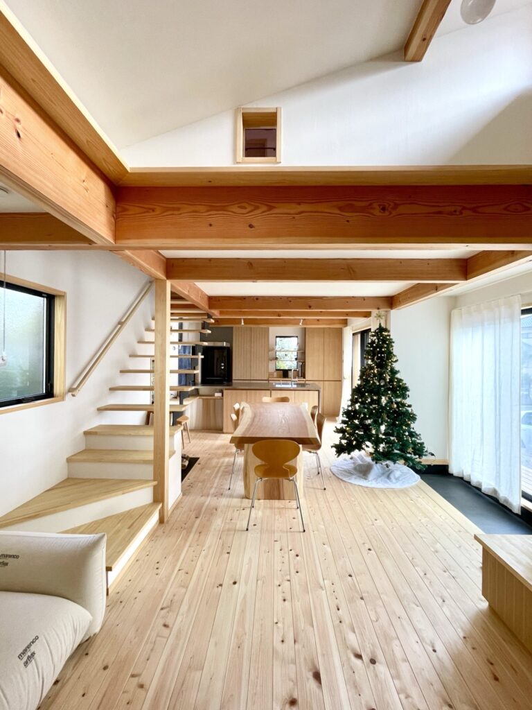 神奈川県鎌倉市西鎌倉に建築したL字型間取り設計の自然素材の家。夏や梅雨時は湿気や熱を排気し、冬は太陽熱で温めた外気を床下に送り換気をしながら床暖房するソーラーシステムそよ風で家中足元から暖かくします。壁天井は消臭調湿効果が高い珪藻土、床は桧フローリングなど天然素材仕上げ。自然素材とソーラーシステムそよ風の相乗効果で湿気が少なく夏涼しく冬暖かいパッシブデザインの環境共生住宅です。 全室収納は造り付けで作成したのでスッキリものが片付き使い勝手と住み心地がいいサステナブルハウス。 画像はウッドデッキに隣接した土間がある吹抜けのリビング。テーブルは杉の一枚もの。椅子はアリンコチェアーとセブンチェアー。ナチュラルインテリアに合う天然素材の麻のカーテンを取り付け。