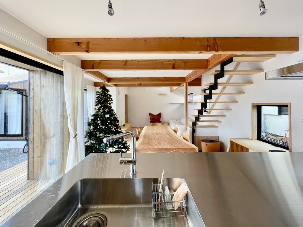 神奈川県鎌倉市西鎌倉に建築したL字型間取り設計の自然素材の家。夏や梅雨時は湿気や熱を排気し、冬は太陽熱で温めた外気を床下に送り換気をしながら床暖房するソーラーシステムそよ風で家中足元から暖かくします。壁天井は消臭調湿効果が高い珪藻土、床は桧フローリングなど天然素材仕上げ。自然素材とソーラーシステムそよ風の相乗効果で湿気が少なく夏涼しく冬暖かいパッシブデザインの環境共生住宅です。 全室収納は造り付けで作成したのでスッキリものが片付き使い勝手と住み心地がいいサステナブルハウス。 画像はウッドデッキに隣接した土間がある吹抜けのリビング。テーブルは杉の一枚もの。