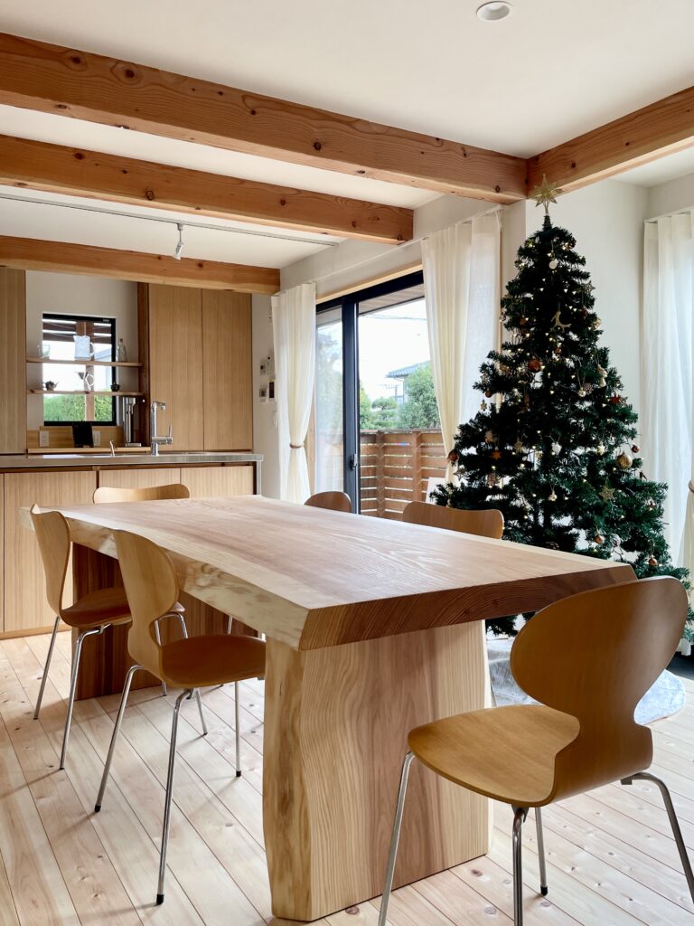 神奈川県鎌倉市西鎌倉に建築したL字型間取り設計の自然素材の家。夏や梅雨時は湿気や熱を排気し、冬は太陽熱で温めた外気を床下に送り換気をしながら床暖房するソーラーシステムそよ風で家中足元から暖かくします。壁天井は消臭調湿効果が高い珪藻土、床は桧フローリングなど天然素材仕上げ。自然素材とソーラーシステムそよ風の相乗効果で湿気が少なく夏涼しく冬暖かいパッシブデザインの環境共生住宅です。 全室収納は造り付けで作成したのでスッキリものが片付き使い勝手と住み心地がいいサステナブルハウス。 画像はウッドデッキに隣接した土間がある吹抜けのリビング。テーブルは杉の一枚もの。椅子はアリンコチェアーとセブンチェアー。ナチュラルインテリアに合う天然素材の麻のカーテンを取り付け。