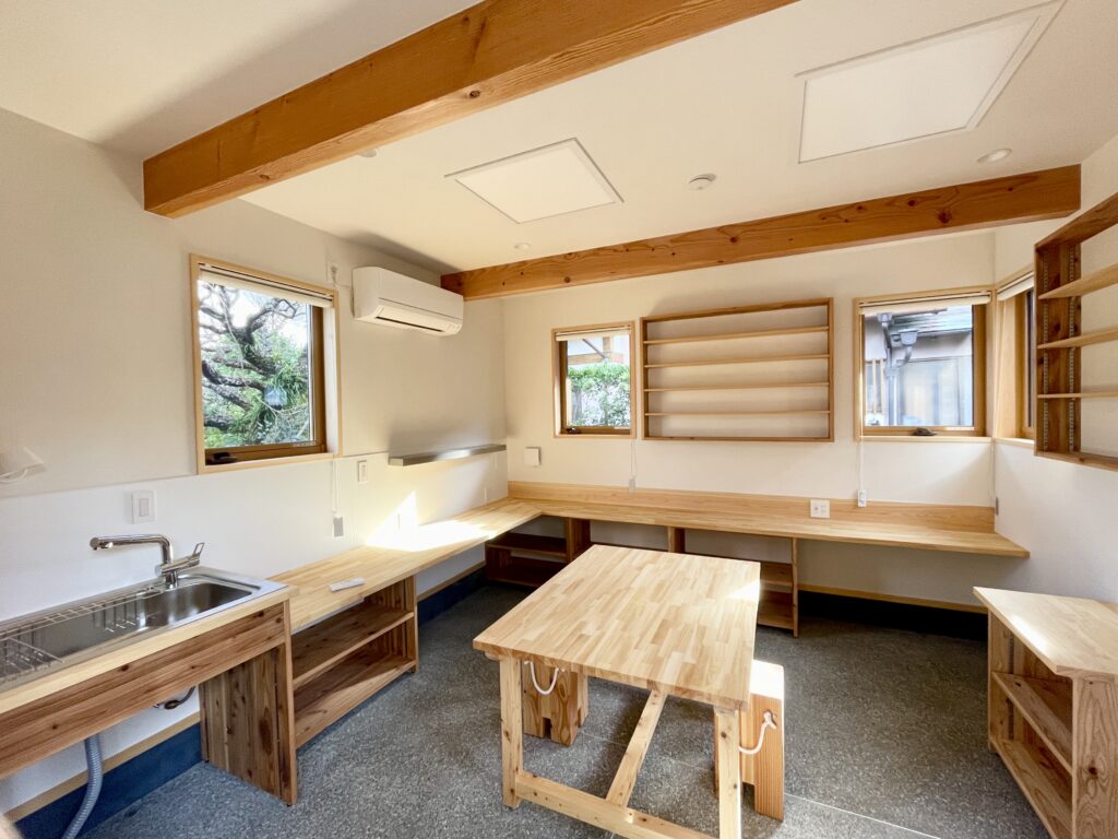 静岡県東部沼津市に建築したコの字型間取り設計の平家の自然素材の家。夏や梅雨時は湿気や熱を排気し、冬は太陽熱で温めた外気を床下に送り換気をしながら床暖房するソーラーシステムそよ風で家中足元から暖かくします。壁天井は消臭調湿効果が高い珪藻土、床は桧フローリングなど天然素材仕上げ。自然素材とソーラーシステムそよ風の相乗効果で湿気が少なく夏涼しく冬暖かいパッシブデザインの環境共生住宅です。 全室収納は造り付けで作成したのでスッキリものが片付き使い勝手と住み心地がいいサステナブルハウス。 画像は玄関土間続きの陶芸アトリエ。ろくろが２台置けるスペースがあり収納棚や展示だなは造り付けで作成。