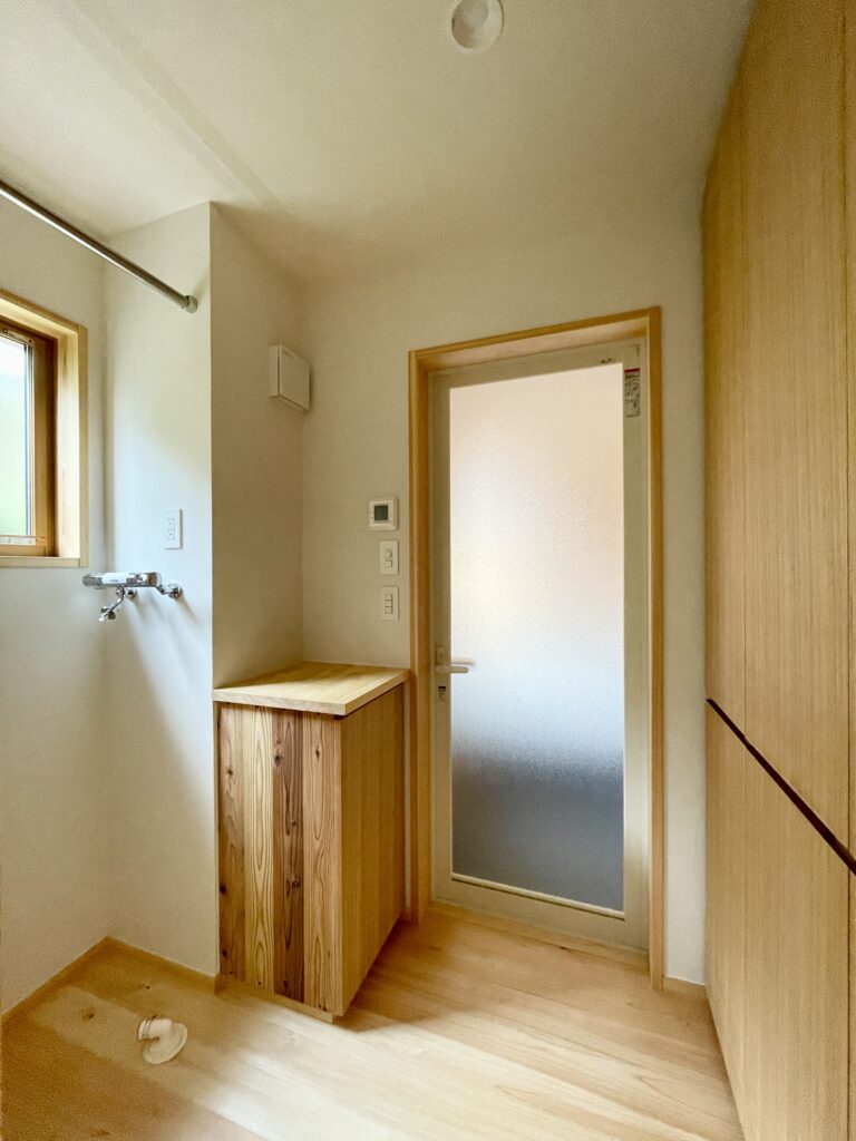 静岡県東部沼津市に建築したコの字型間取りの自然素材の家 。梅雨時や夏は熱気や湿気を排気し冬は太陽熱で温めた外気を床下に送り換気をしながら床暖房するソーラーシステムそよ風で家中足元から暖かい環境共生住宅です。 全室壁天井は消臭調湿効果が高い珪藻土床は桧のフローリングなど天然素材仕上げ 。自然素材とソーラーシステムそよ風の相乗効果で湿気が少なく夏涼しく冬暖かい明るく風通しのいいパッシブデザインの注文住宅 。全室収納を造り付けで作成しスッキリものが片付き使い勝手と居心地がいいサステナブルハウス 。画像はバスタオルや下着がしまえる収納棚がある脱衣室。洗濯物仮干し用のパイプも設置。
